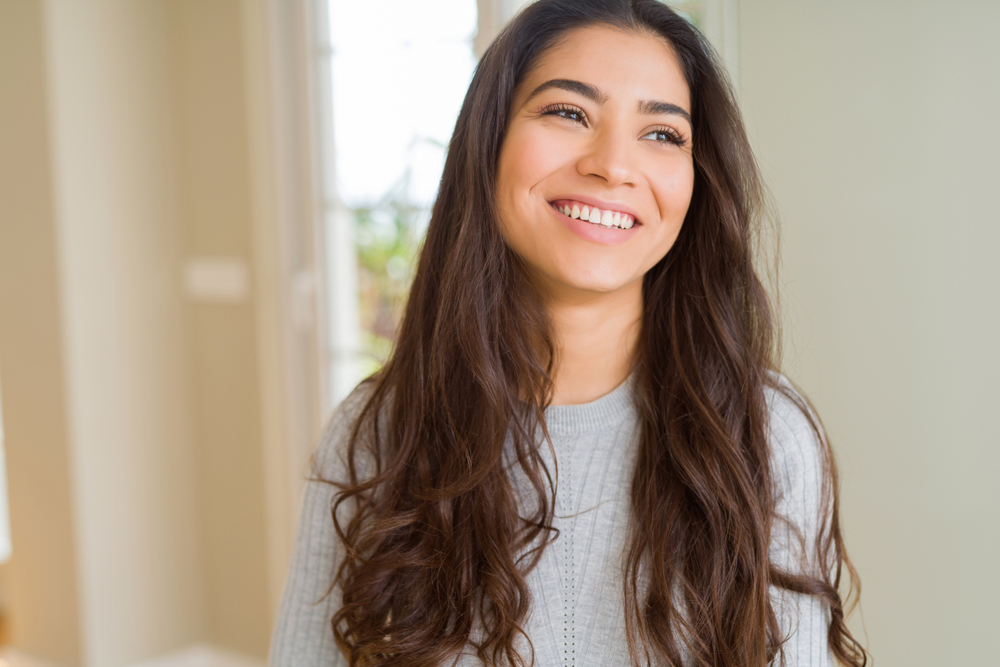 Smiling young woman with long hair indoors, wearing gray sweater, radiating natural confidence