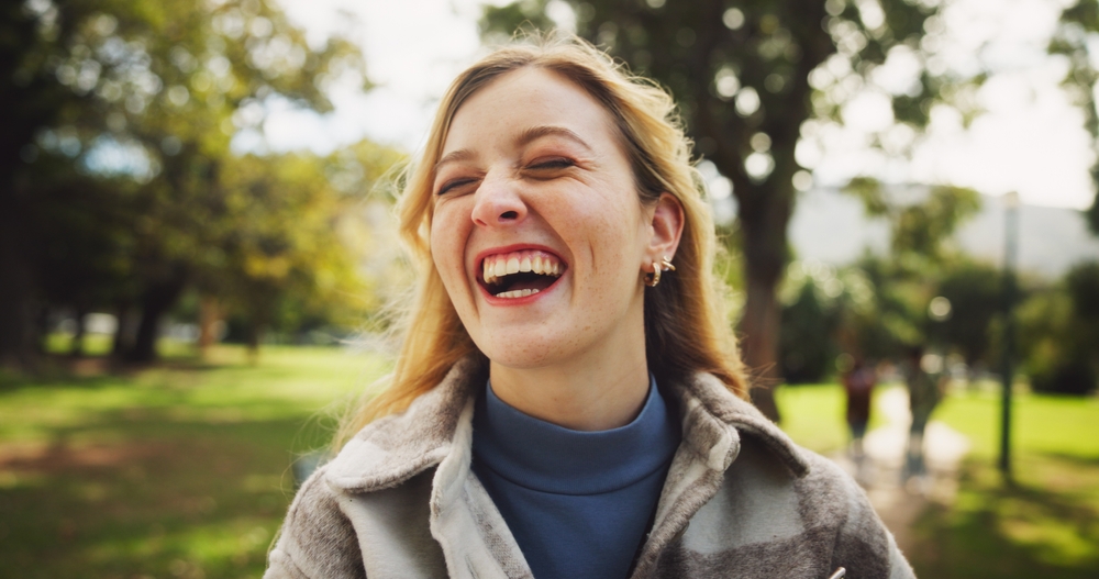 A young woman laughing joyfully outdoors, wearing a cozy coat in the park.